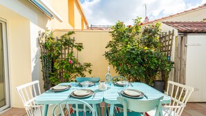A turquoise table set with plates, glasses, and cutlery, surrounded by white chairs, in a sunny outdoor patio area with potted plants and a tiled floor. - Maison au cœur de la Chaume + Garage fermé (Les Sables-d'Olonne)