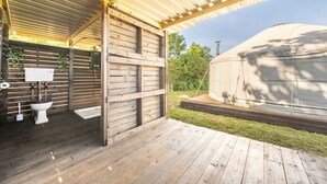 Un baño exterior cubierto con inodoro y lavamanos, una terraza de madera y una tienda de campaña grande al fondo.
