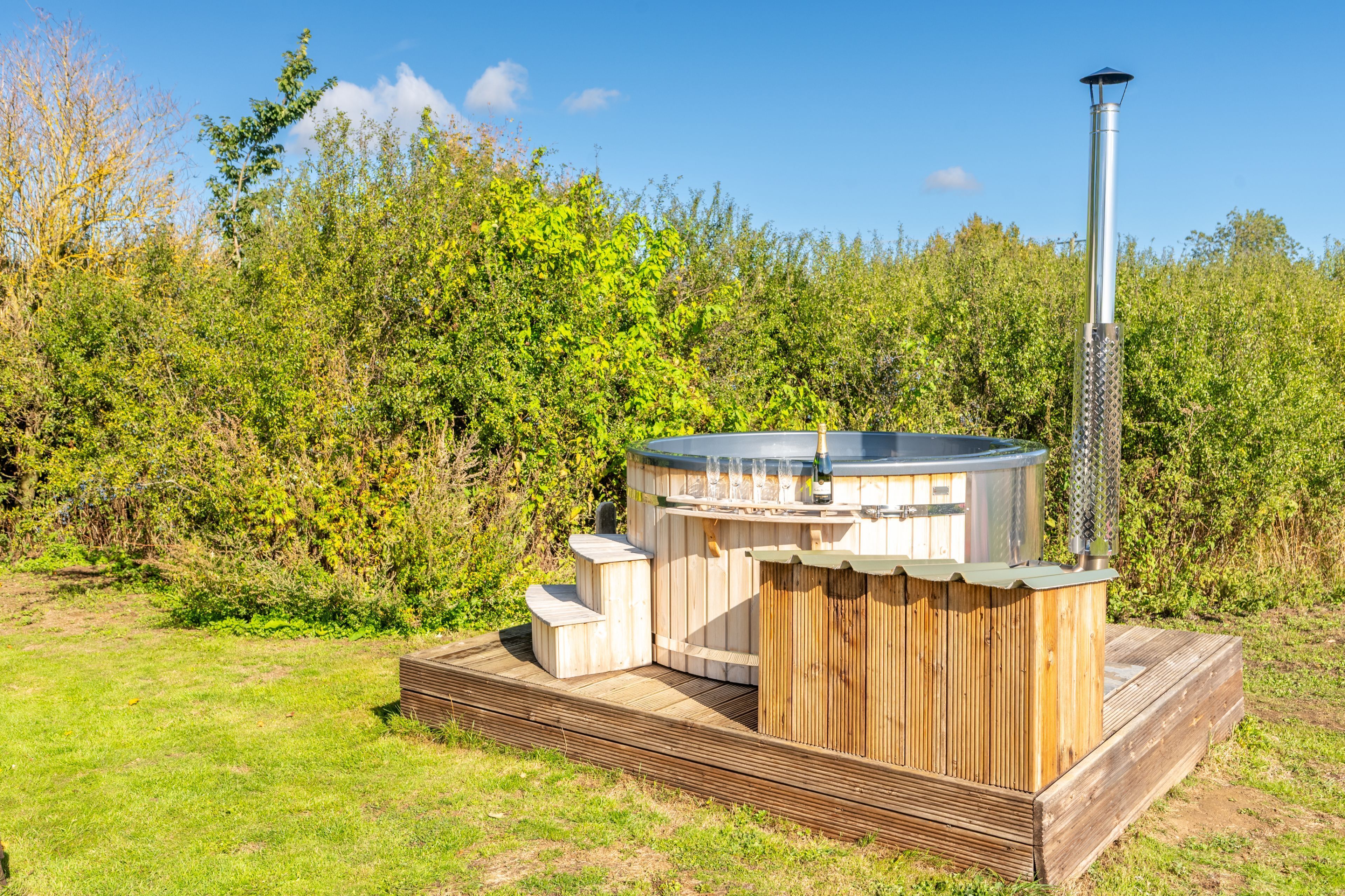 Een hottub met een houten terras en schoorsteen, omgeven door groen.