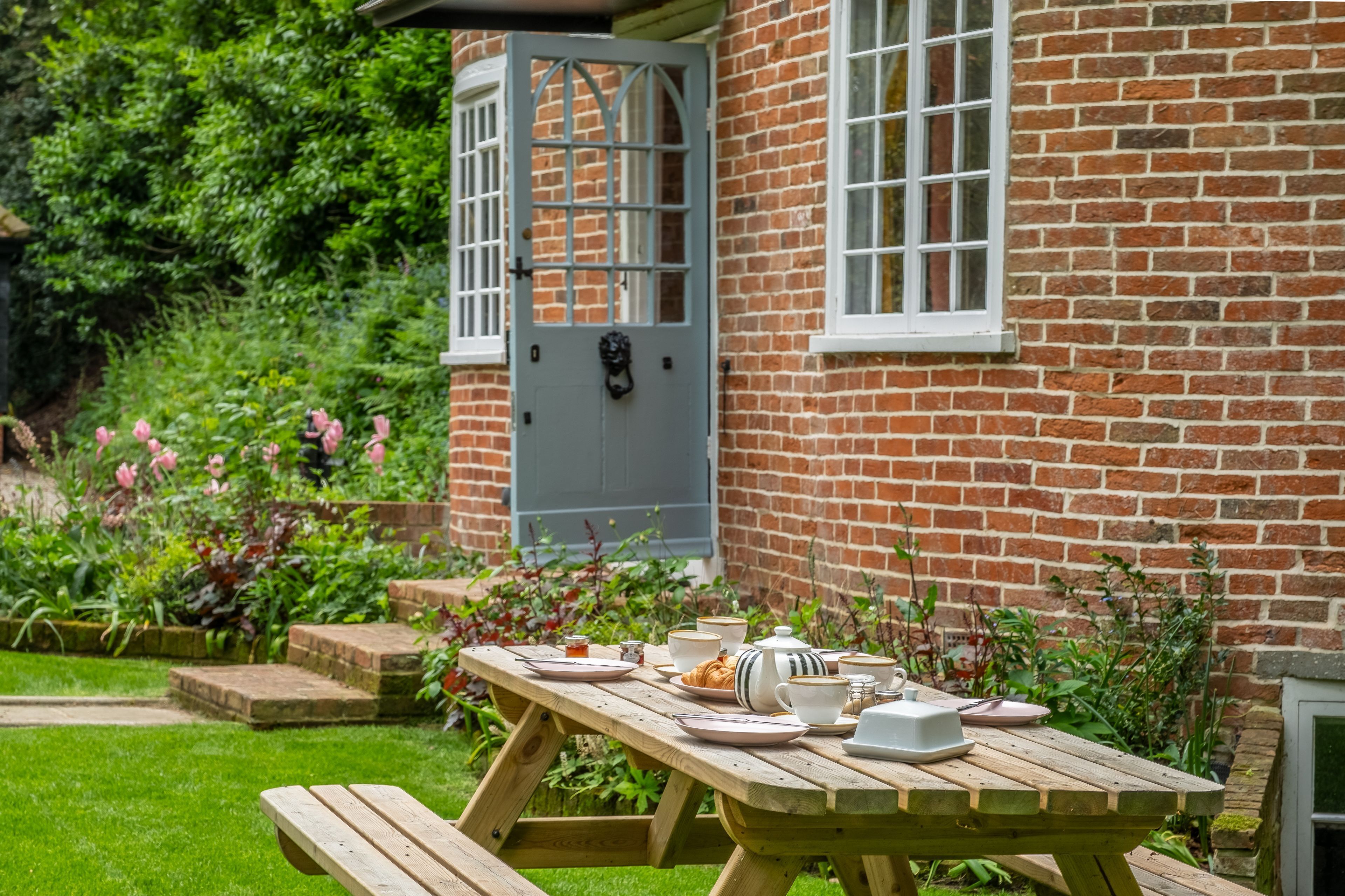 Een houten picknicktafel in een tuin, met een bakstenen huis op de achtergrond.