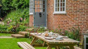 A wooden picnic table set up in a garden with a brick house in the background. - A fairy-tale luxurious cottage - The Tea Caddy (Colchester)