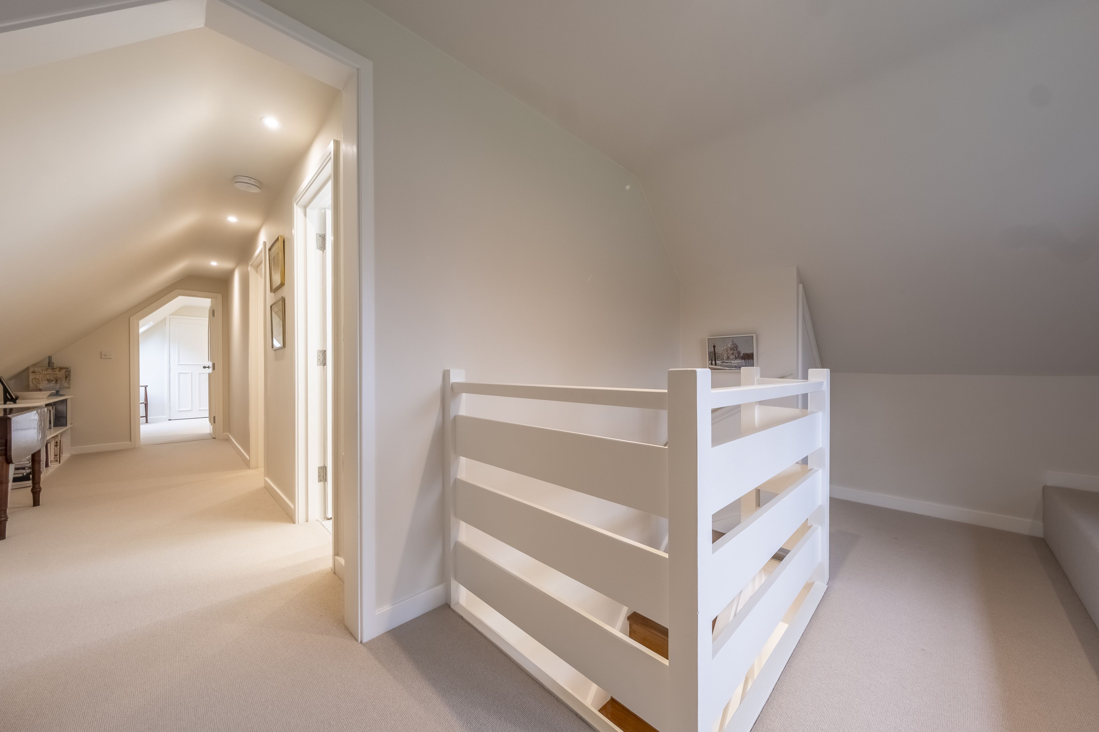 Un espacioso dormitorio en el ático con techo inclinado, estructura de cama blanca y vista al pasillo.