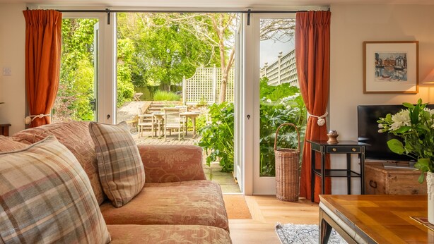 A cozy living room with a sofa, a TV, and a view of a garden through glass doors. - Barn conversion with private pool - Yew Tree Barn (Colchester)
