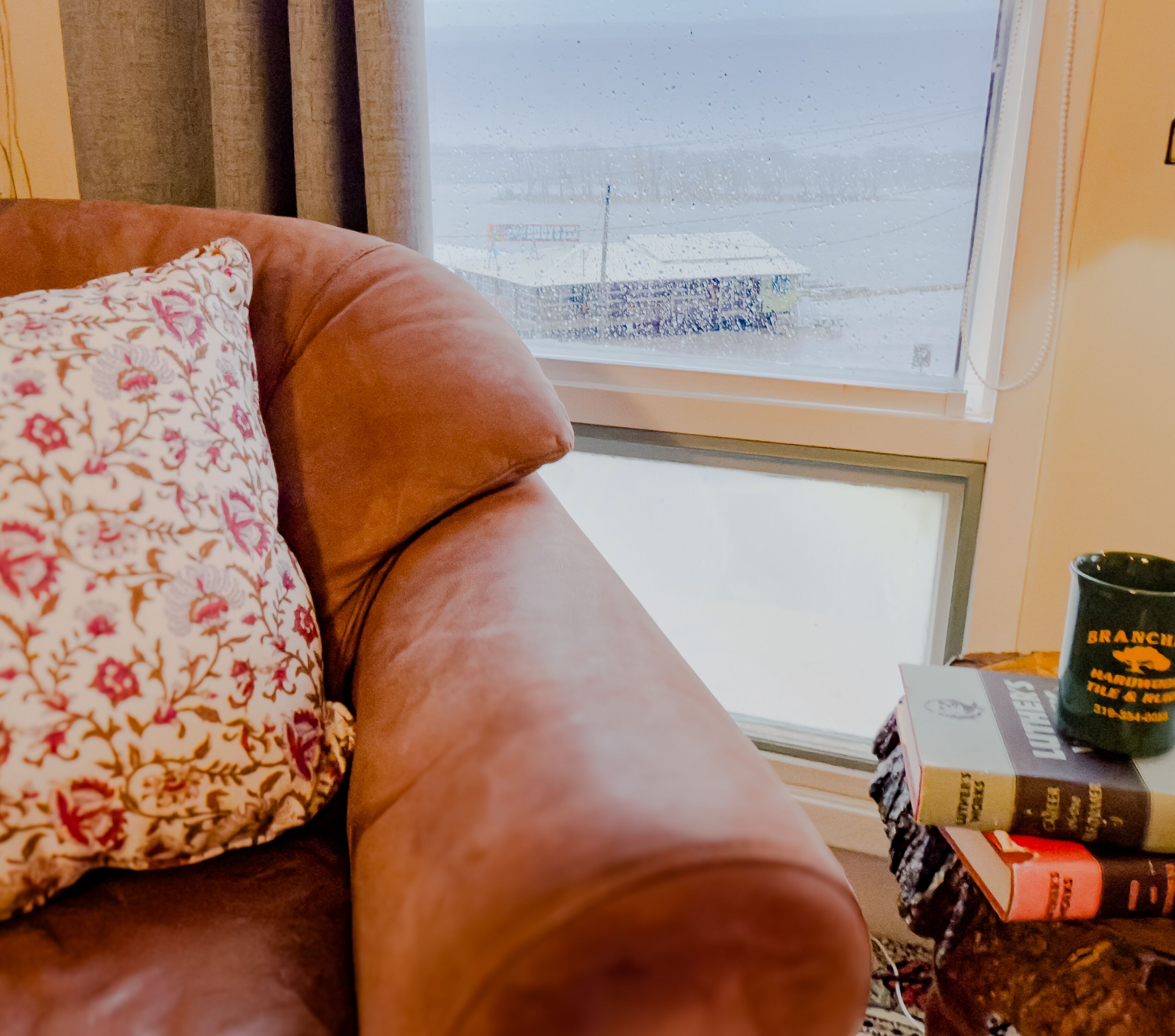 A brown leather sofa with a floral pillow, a window with a view of a snowy landscape, and a stack of books and a mug on the windowsill.