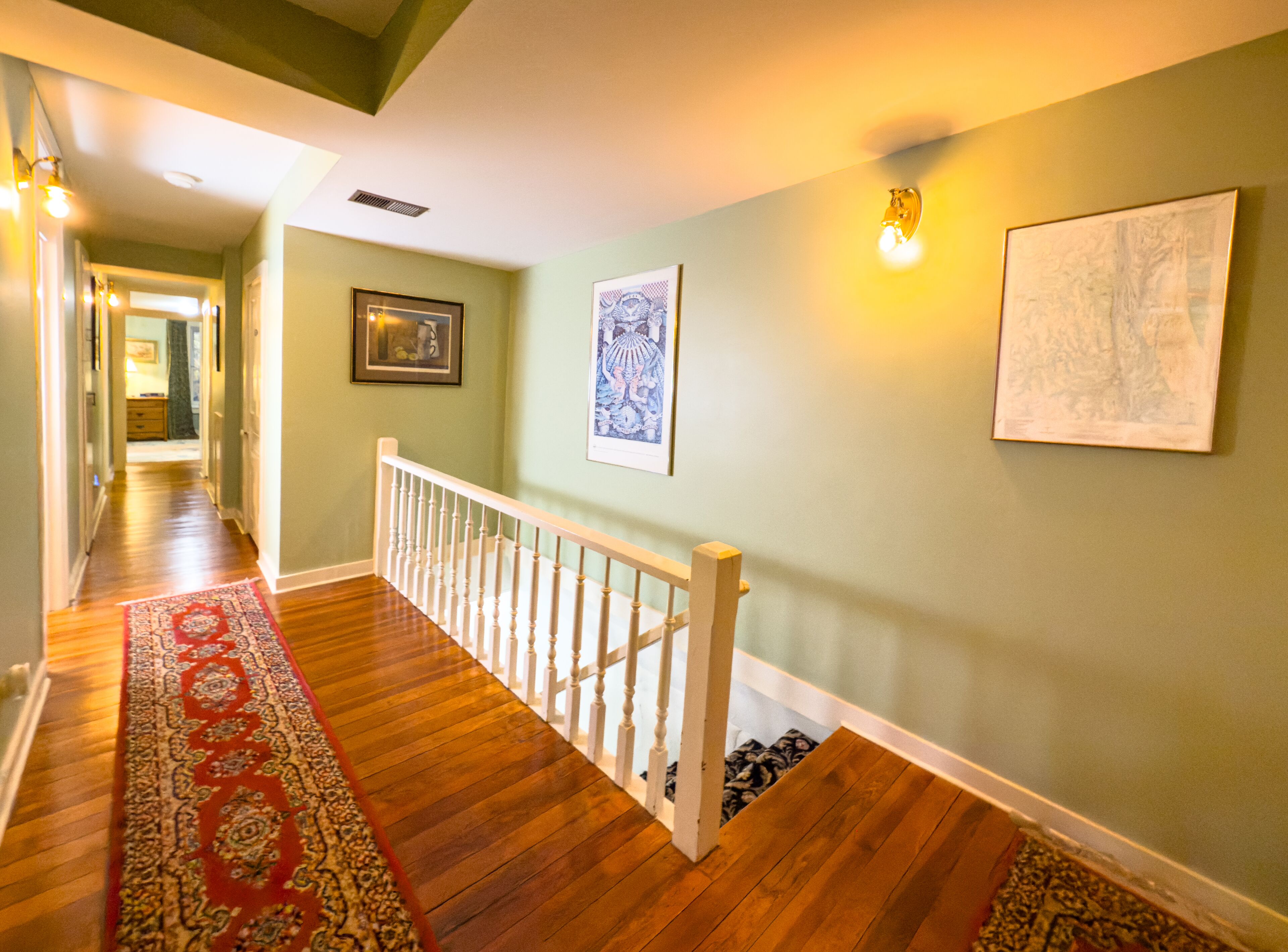 A hallway with wooden flooring, a white railing, and artwork on the walls.