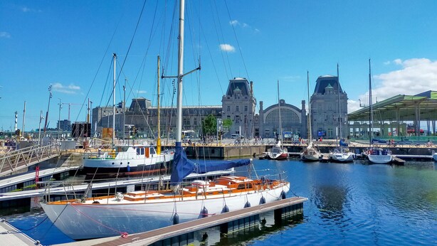 Un port de plaisance avec des bateaux amarrés, un grand bâtiment doté d’une tour horloge et une promenade couverte.