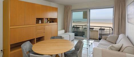 A modern living room with a wooden cabinet, a round dining table, grey upholstered chairs, a beige sofa, and a balcony with a view of the beach.