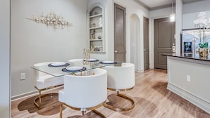 Un comedor moderno con una mesa de vidrio y sillas blancas, un adorno en la pared y una cocina al fondo.