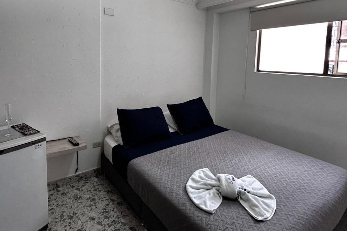 A hotel room with a bed, a small bedside table with a remote control, a glass of water, and a white appliance.