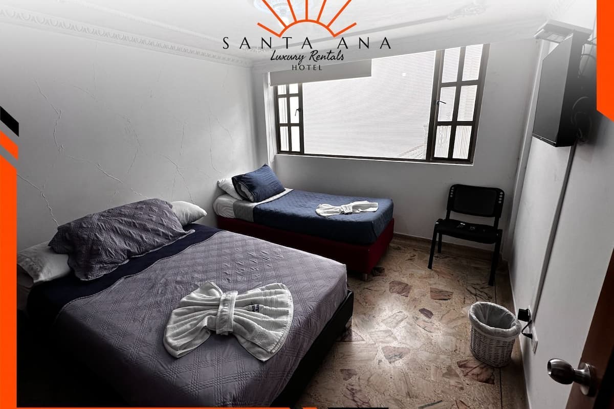 A hotel room with two beds, a window with blinds, a chair, and a trash bin.