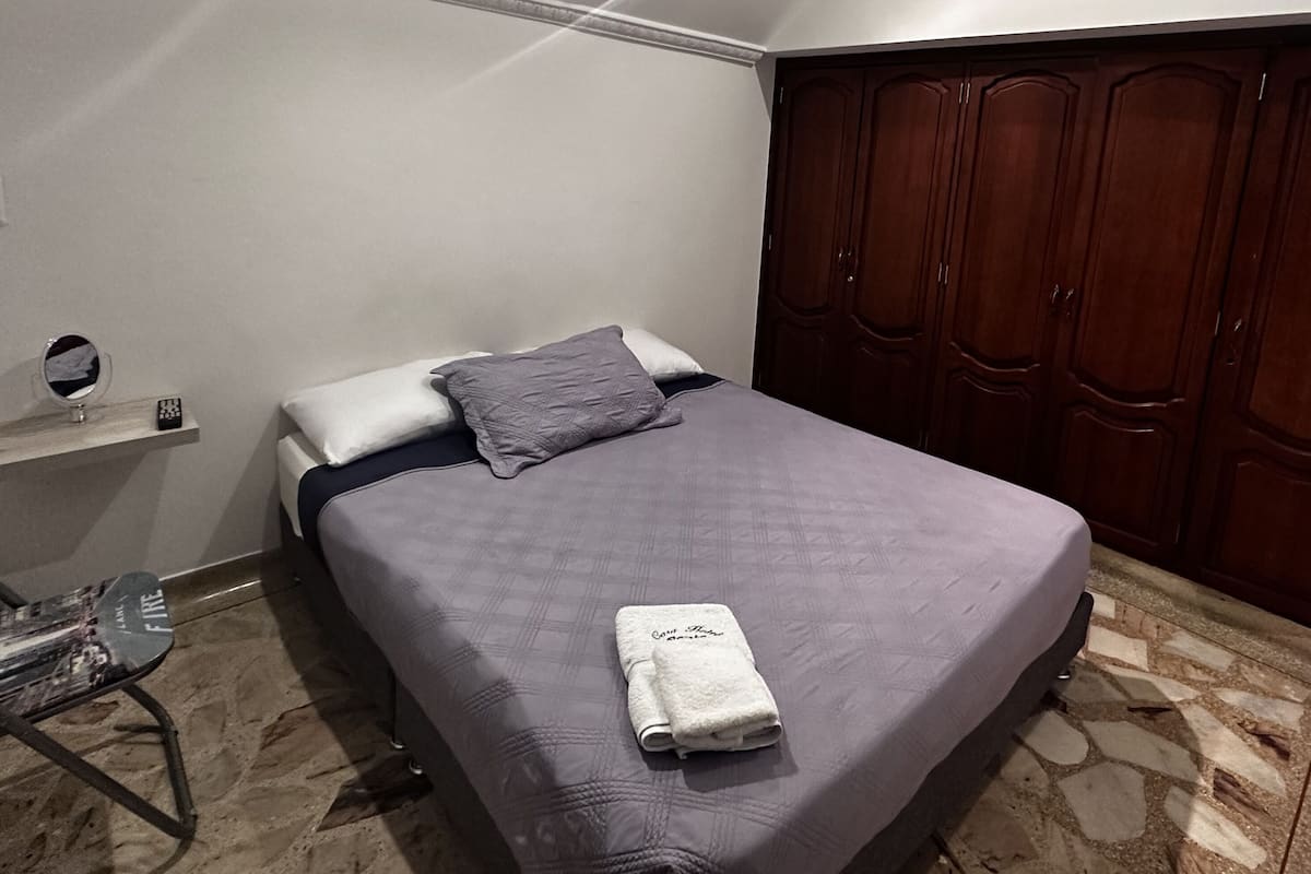 A bedroom with a bed, a wooden wardrobe, a small bedside table with a mirror, and a chair.