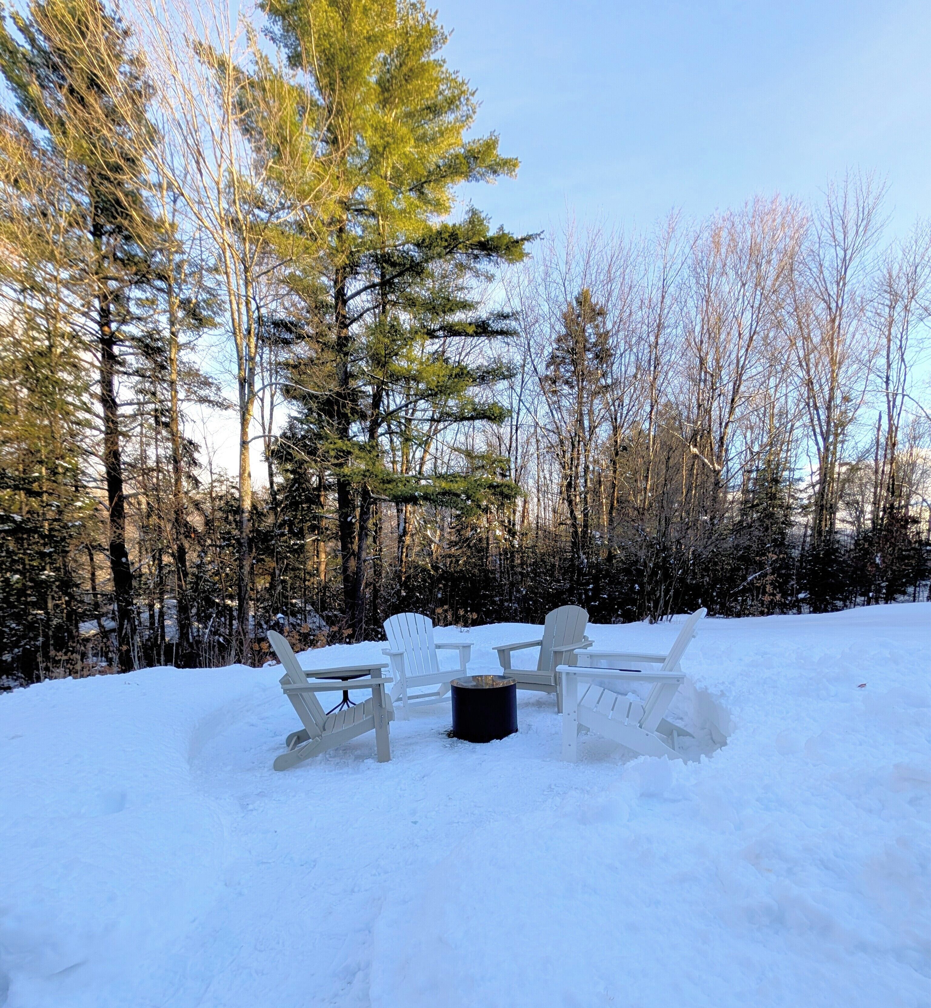 A snow-covered outdoor seating area with Adirondack chairs and a fire pit.