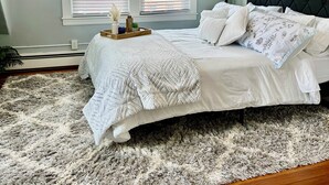 A bedroom with a bed, a ceiling fan, two windows with blinds, a nightstand, a lamp, and a painting on the wall.