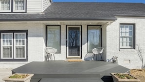 Een tweelaags huis met een overdekte veranda, twee witte stoelen en een kleine tuin.
