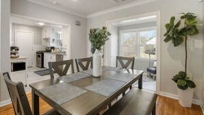 Een eetgedeelte met een lange tafel, stoelen en een vaas met bloemen. Achtergrond is een keuken met kastjes en een raam.