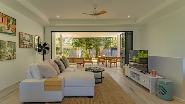 A modern living room with a white sofa, a TV, and a fan. There is a dining area with a table and chairs visible through an open doorway. - Escape to Serenity: Your Dreamy 4-BR Mountain View Getaway (Tambon Rawai)