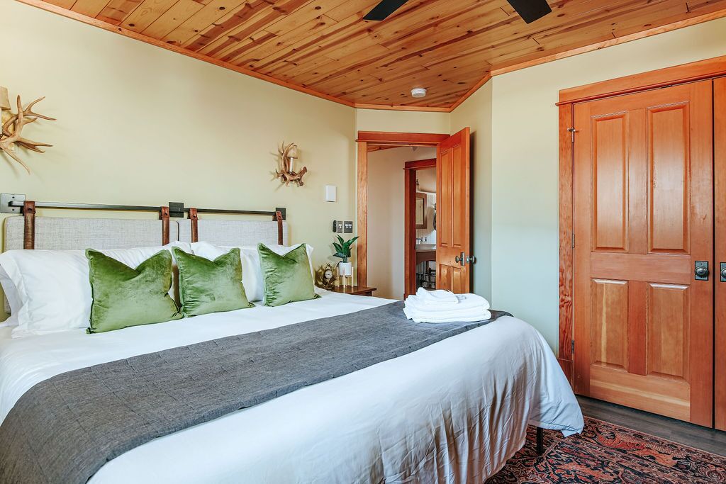 A neatly made bed with a grey blanket and green pillows, a wooden ceiling, and a wooden door.