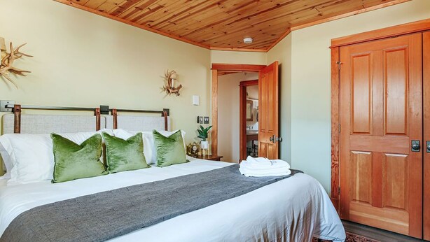 Ein ordentlich bezogenes Bett mit grauer Decke und grünen Kissen, einer Holzbalkendecke und einer Holztür.