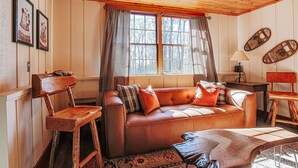 Ein gemütlicher Raum mit einem braunen Ledersofa, Holzstühlen, einem gemusterten Teppich und einem großen Fenster mit Vorhängen.