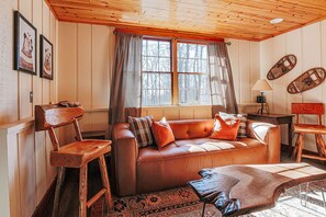 Ein gemütlicher Raum mit einem braunen Ledersofa, Holzstühlen, einem gemusterten Teppich und einem großen Fenster mit Vorhängen.