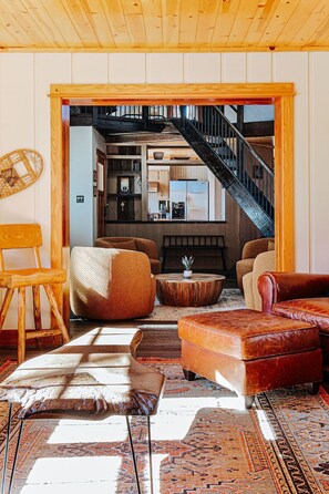 A cozy living room with a wooden ceiling, a staircase leading to an upper level, and a view of a kitchen and dining area.