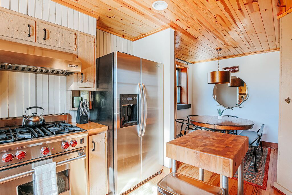 A modern kitchen with a stainless steel refrigerator, a gas stove, wooden cabinets, and a wooden dining table.