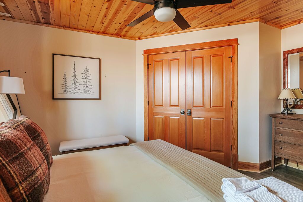 A bedroom with a bed, a wooden wardrobe, a ceiling fan, a framed picture, and a bedside table with a lamp.