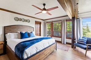 Ein Schlafzimmer mit einem großen Bett, einem Deckenventilator, einem Holzkopfende und Blick auf Grün hinter den Fenstern.