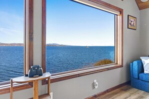 A room with a large window overlooking a body of water, a small table with luggage, and a blue sofa.