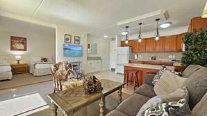 A spacious living room with a sofa, a coffee table, and a TV. There is a kitchen area with wooden cabinets and a refrigerator.