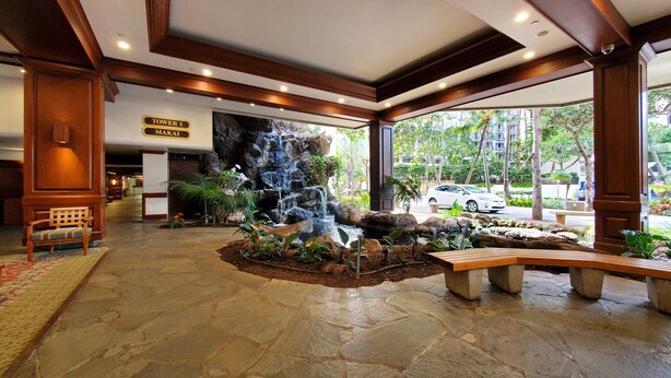 A lobby with a waterfall feature, stone flooring, wooden benches, and a sign indicating 'Tower I Makai'.