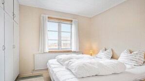 Un dormitorio con cama, cortinas blancas, una ventana, un radiador y una lámpara.