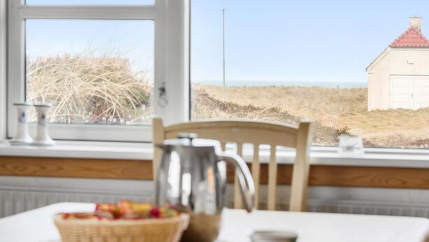 Un comedor con una mesa, sillas, una cesta de fruta, una jarrón y vistas a la playa y un edificio a lo lejos.
