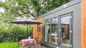 Un espace repas extérieur moderne, avec une table dressée pour deux personnes, un grand parasol et une vue sur la pièce adjacente.