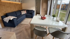 Een moderne woonkamer met een donkerblauwe modulaire bank, een eettafel met glazen blad en twee stoelen, en uitzicht op het buitenleven door grote ramen.