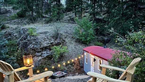 A rustic outdoor seating area with two wooden Adirondack chairs, a small table, and a lantern. A red-roofed cabin is visible in the background. - Runaway Express Coach (Oliver)