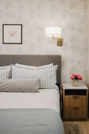 A neatly made bed with pillows, a bedside lamp, a framed picture, and a small wooden nightstand with a vase of flowers. - Luxury Direct Oceanview King Villa • Steps to Beach • Gourmet Kitchen (Hilton Head Island)