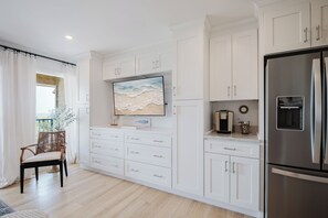 A modern kitchen with white cabinetry, a stainless steel refrigerator, and a flat-screen TV mounted above the counter. - Luxury Direct Oceanview King Villa • Steps to Beach • Gourmet Kitchen (Hilton Head Island)