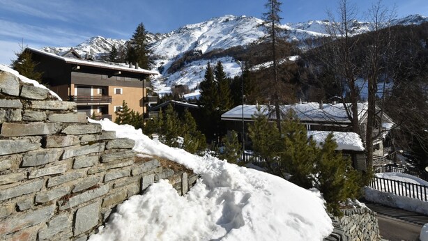Un chalet de montagne aux murs de pierre et au toit enneigé, entouré d’arbres et de montagnes couverts de neige.