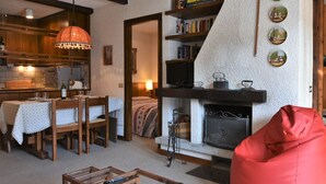 Un salon chaleureux avec une cheminée, un coin repas avec une table et des chaises, et une cuisine dotée de meubles en bois et d’un luminaire suspendu.