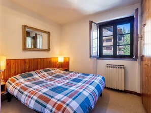 A bedroom with a wooden headboard, a large bed with a colorful quilt, a window with a view of buildings, a radiator, and a mirror on the wall.