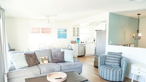 A modern living room with a grey sofa, a round coffee table, and a striped armchair. A kitchen with white cabinets and a refrigerator is visible in the background. - Walk to Beach! Entire SC Triplex (San Clemente)