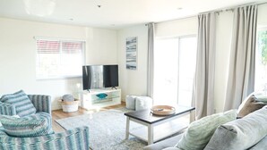 A modern living room with a sofa, armchair, and a coffee table. There is a flat-screen TV on a stand, a window with blinds, and a painting on the wall. - Walk to Beach! Entire SC Triplex (San Clemente)