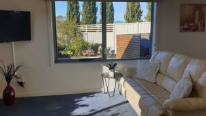Een woonkamer met een beige bank, een klein rond tafeltje, een aan de muur bevestigde televisie en een raam met uitzicht op een hek en bomen.