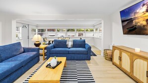 Una sala de estar moderna con un sofá azul, una mesa de centro de madera y un televisor grande mostrando una escena de atardecer.