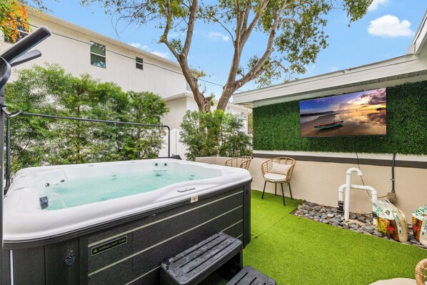 Un bain à remous avec vue sur le coucher de soleil, un téléviseur fixé au mur et un petit espace jardin aménagé avec du gazon synthétique et des rochers.