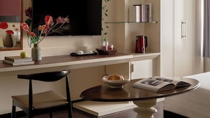 Une chambre d’hôtel équipée d’un téléviseur à écran plat fixé au mur, d’un bureau avec une chaise, d’une petite table avec un bol et un magazine, et d’un lit recouvert d’une couette blanche.