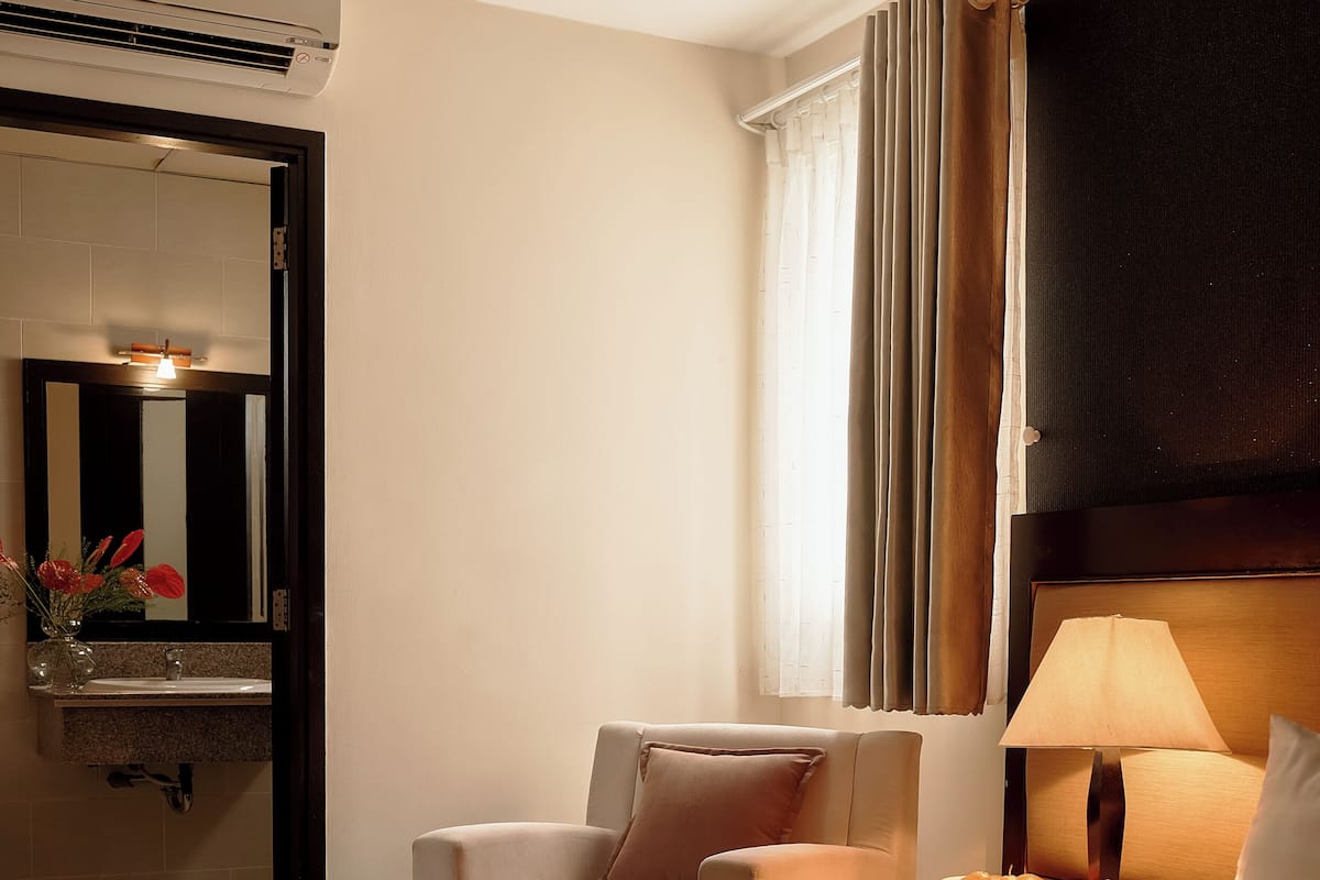 Une chambre d’hôtel comprenant un lit, une chaise, une table de chevet avec une lampe, un miroir et un appareil de climatisation.