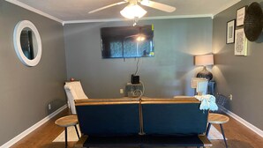 A living room with a dark blue sofa, a ceiling fan, a flat-screen TV, a round mirror, and a small side table. - Beautiful 3Beds in Bridge City! (Bridge City)