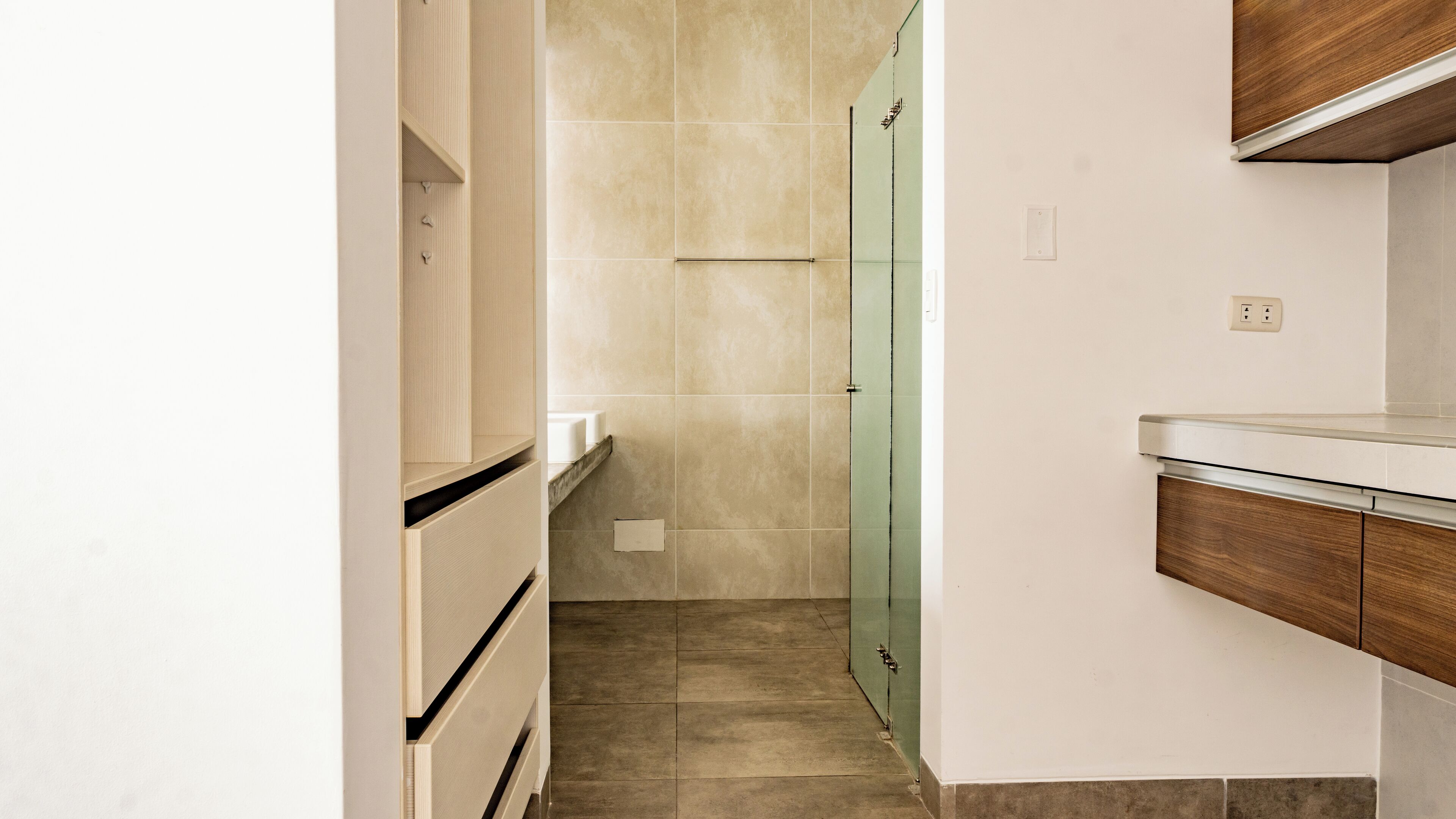 A narrow hallway with a glass door leading to a bathroom, a wooden cabinet on the right, and a white countertop on the left.